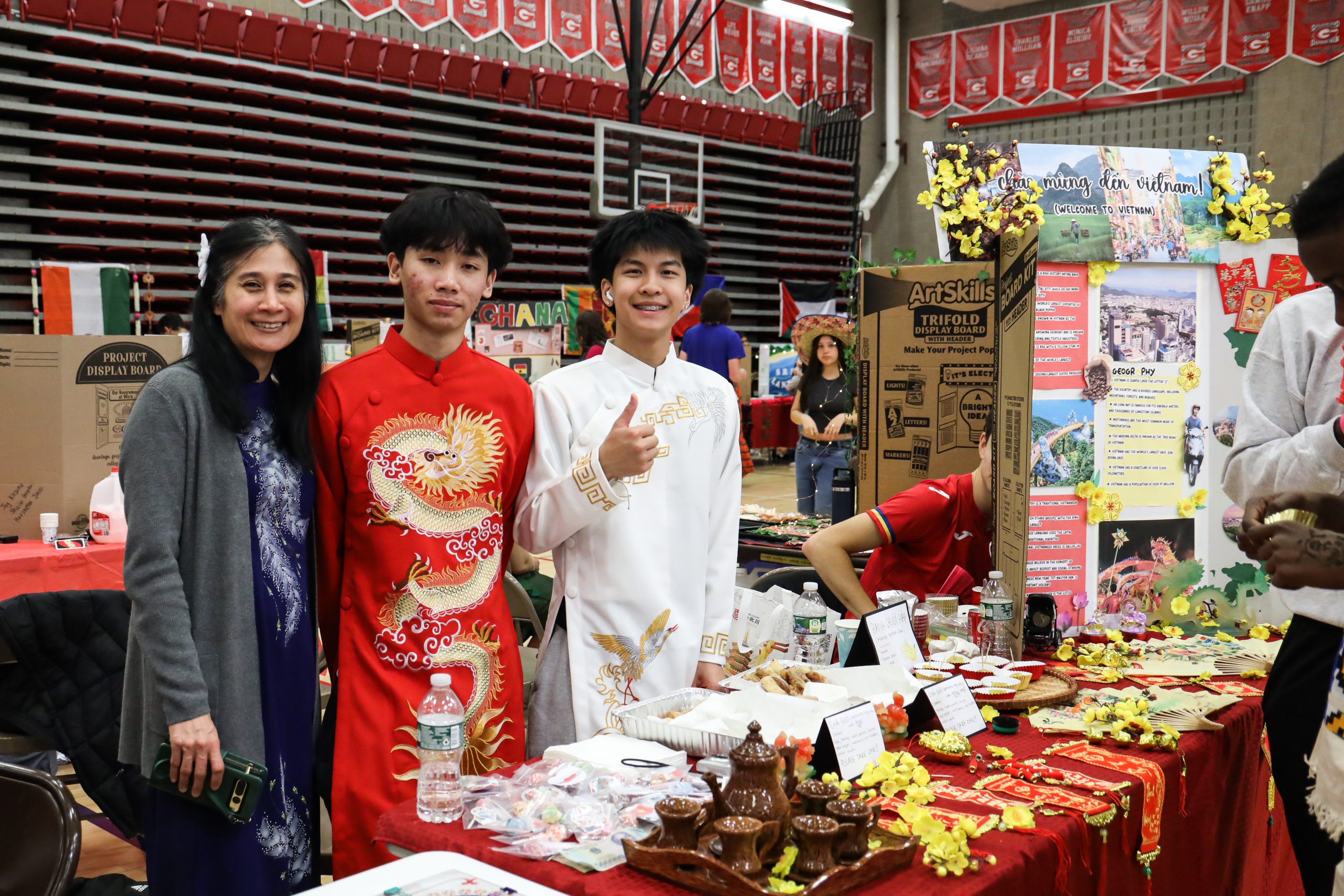 Two students and an adult stand behind a table that has a red tablecloth and a tea set. The students are wearing traditional Chinese clothing. One student is giving a thumbs up
