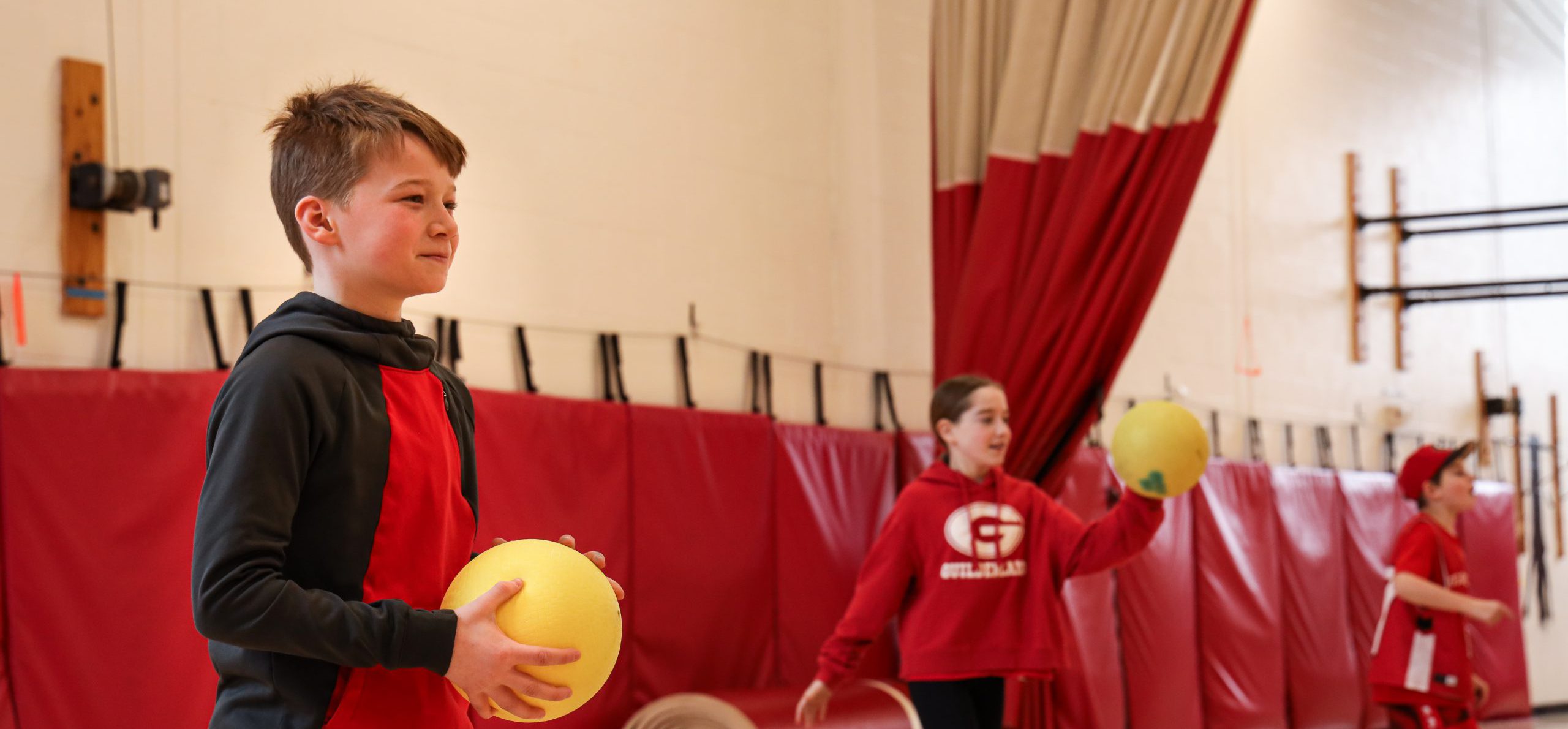 Elementary student in P.E. class. The center student is holding a ball and the student in the background is throwing a ball.