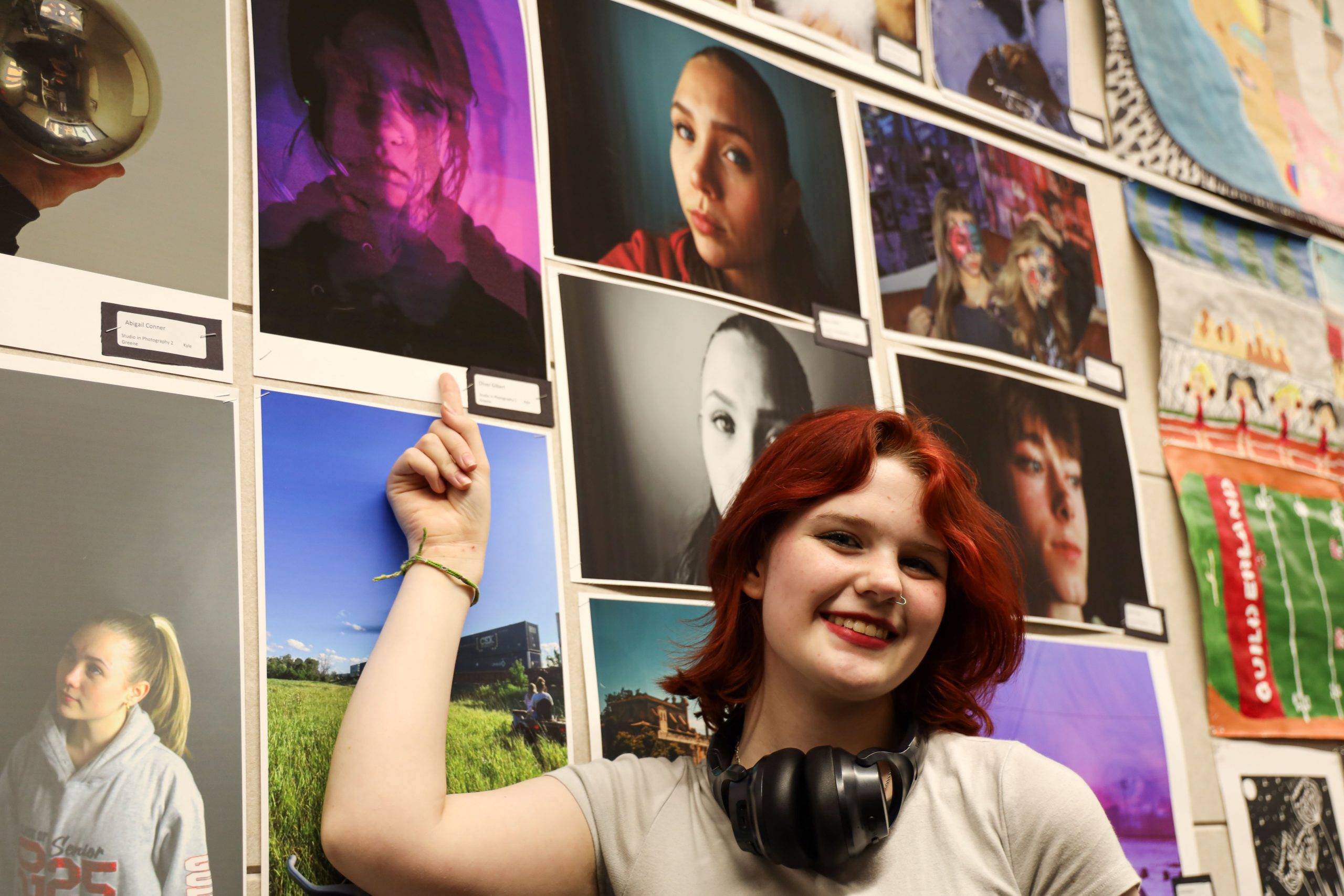 A high school student wearing large headphones around her neck is standing in front of a wall that has a lot of student artwork posted. She is pointing to a piece of artwork. Her t-shirt is light tan and her hair is red
