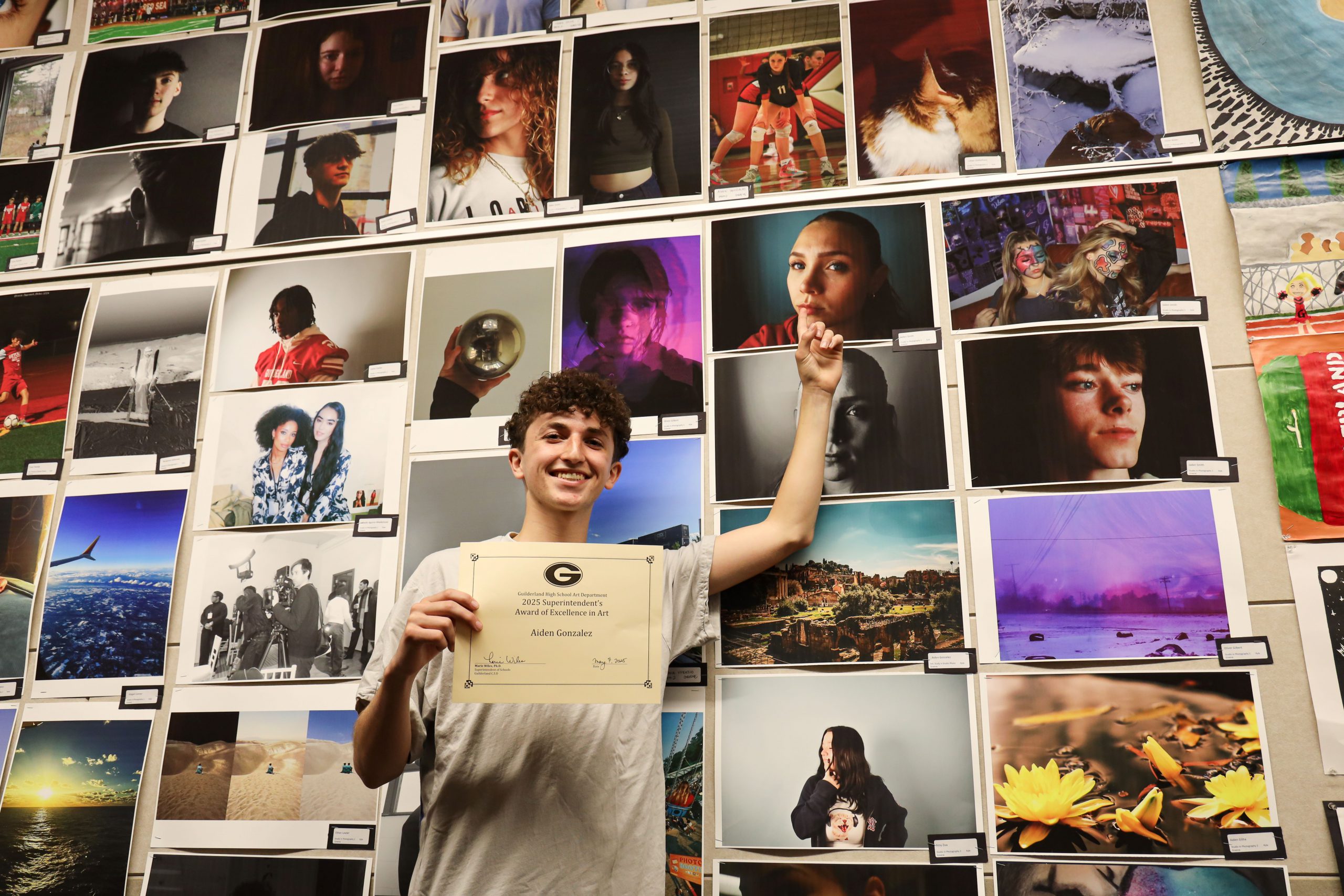 A high school student is standing in front of a wall with a lot of student artwork hanging on it. You can see many pieces. He is holding a certificate and is pointing to a piece of artwork and smiling