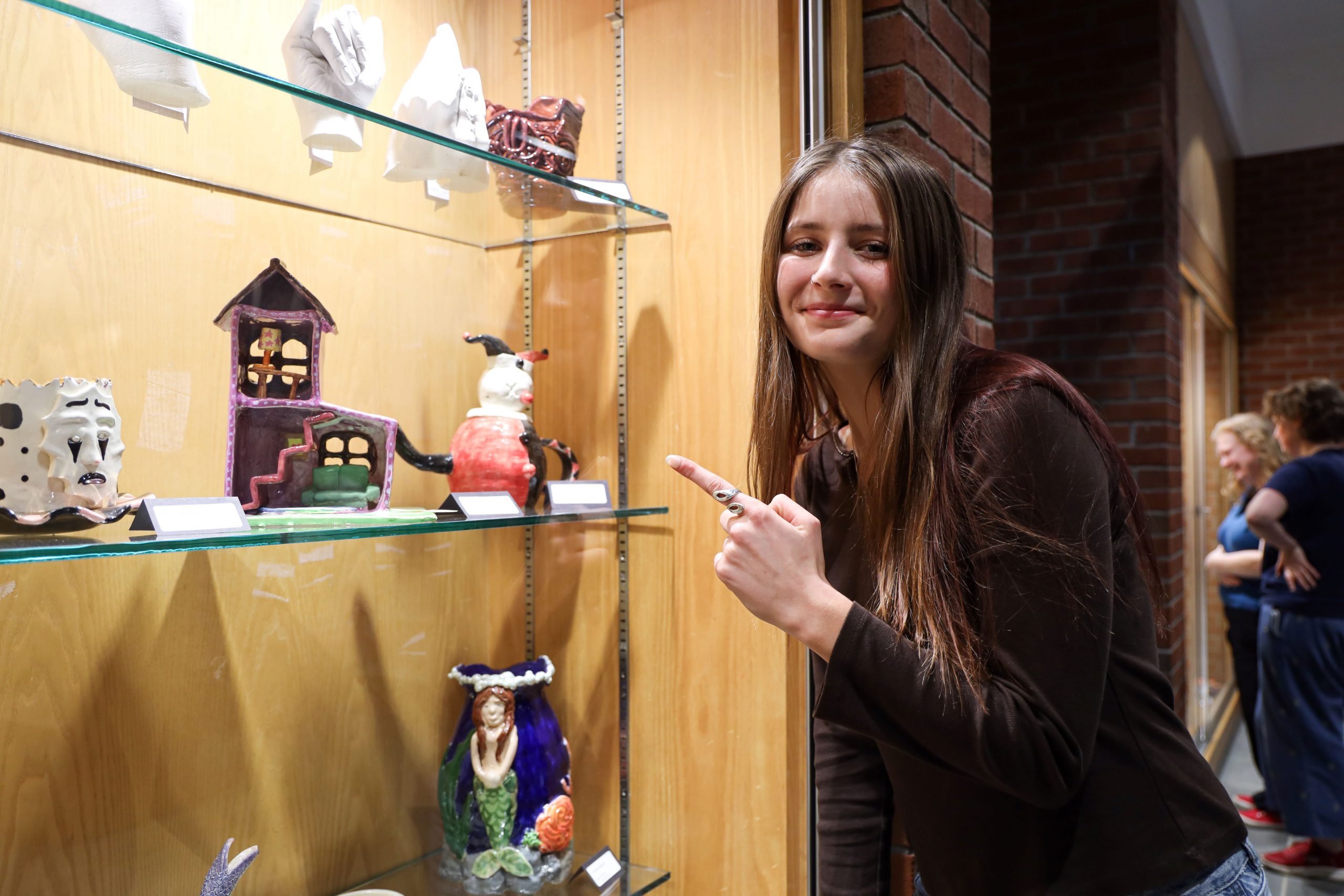 A high school student is pointing to a piece of artwork in a display case. She has long hair and is smiling