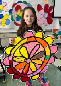 A young student holds a colorful piece of artwork and is smiling. She is standing in a classroom; behind her is colorful artwork.