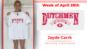 A student stands in front of a white wall, holding a white t-shirt with red lettering that reads "Dutchmen of the Week."