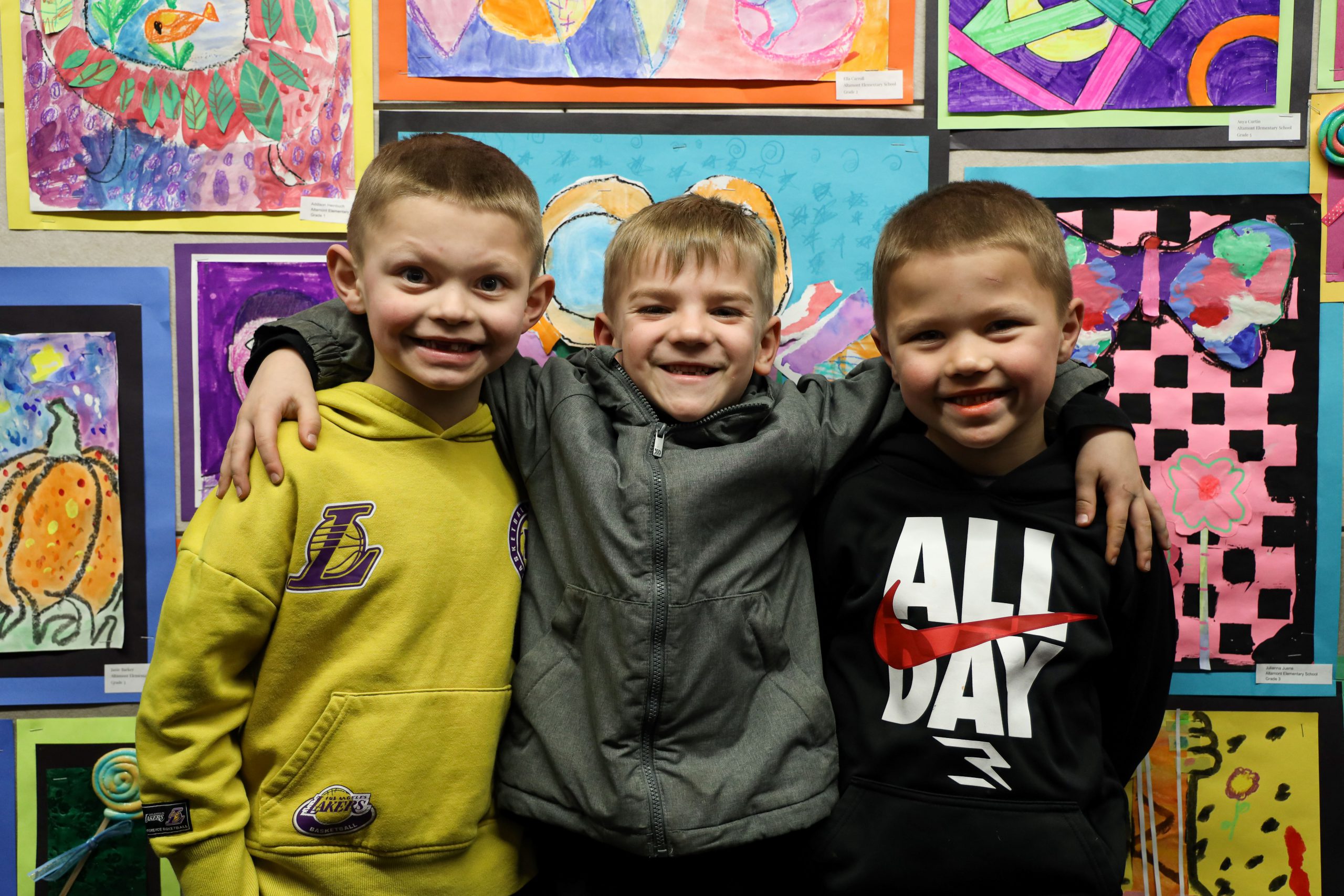 Three young boys are standing in a row, their arms are around each other. They are standing in front of student artwork