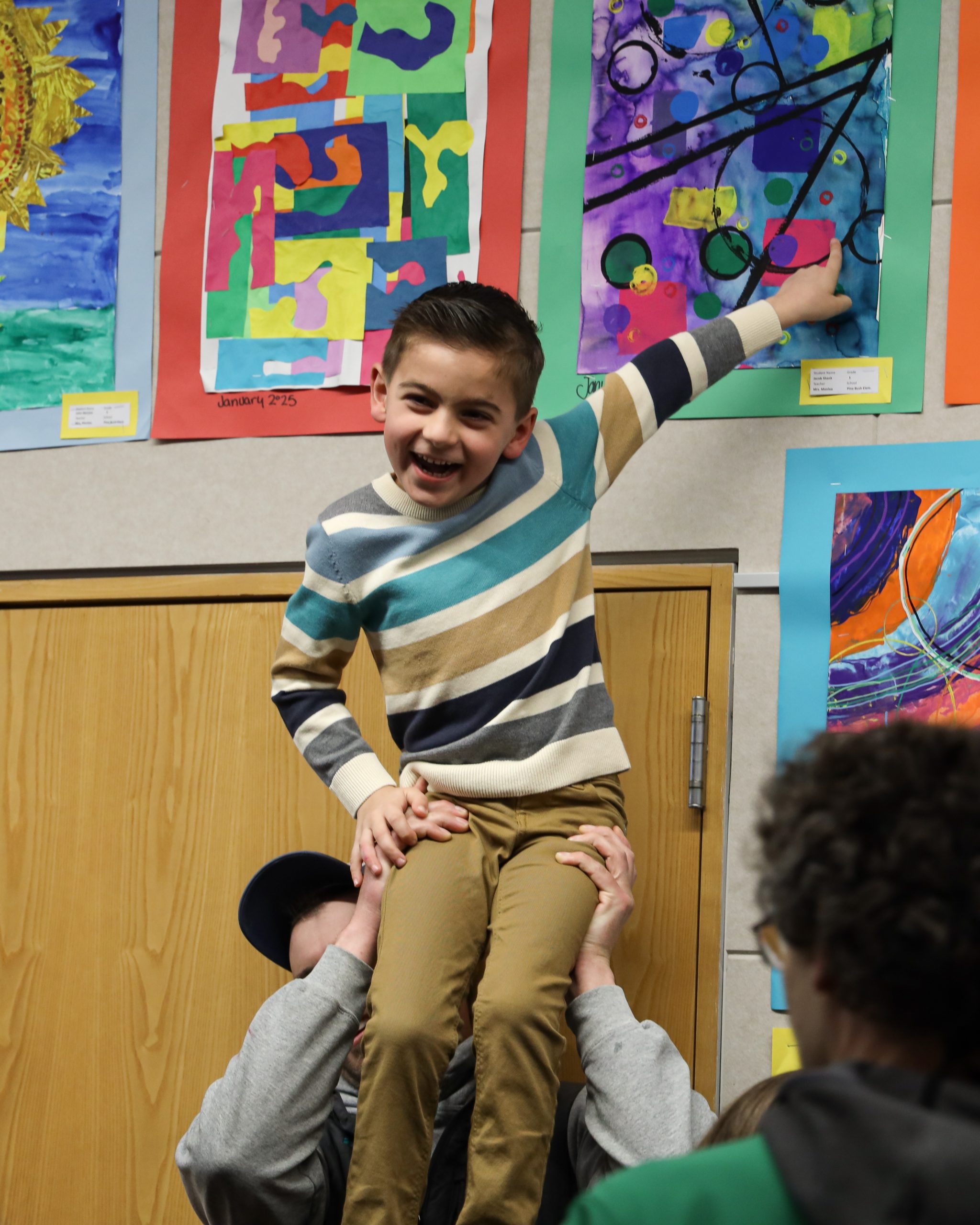 A small child is sitting on a man's child. The boy is pointing at a piece of artwork posted on the wall. The wall is covered with student artwork