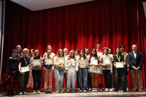 A group of students and two teachers stand on a stage. There is a red curtain in the background. The students are holding certificates and wearing medals