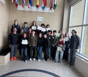 A group of students and teachers stand together at the end of a hallway. They are smiling and looking at the camera. A few are holding up certificates