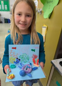 A young student is in a classroom, holding a piece of artwork.