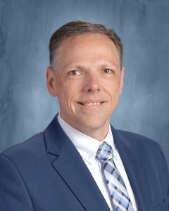 In a headshot, a man, wearing a blue suit, white shirt, blue plaid tie, sitting in front of a blue background, smiles at the camera.