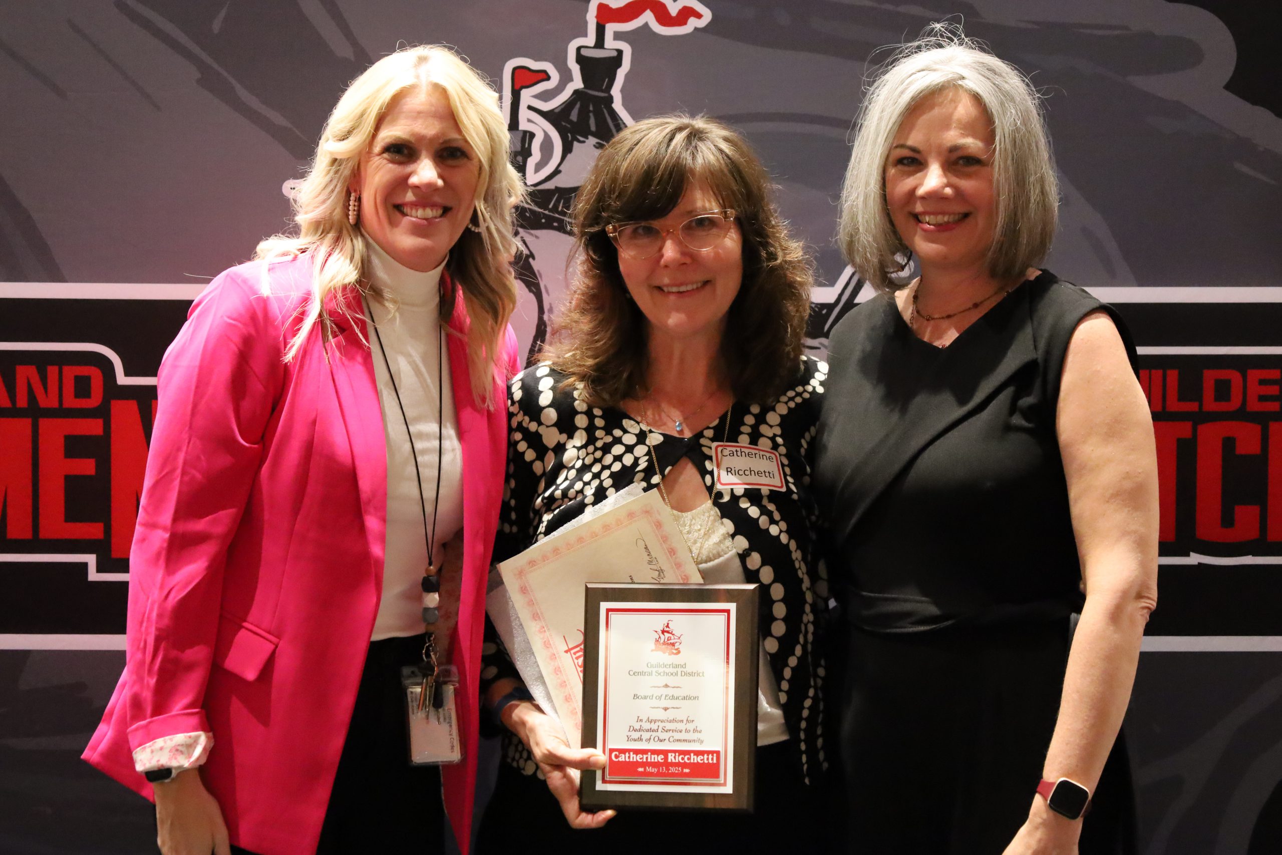Three people stand in front of a backdrop. One is holding a plaque, both are smiling