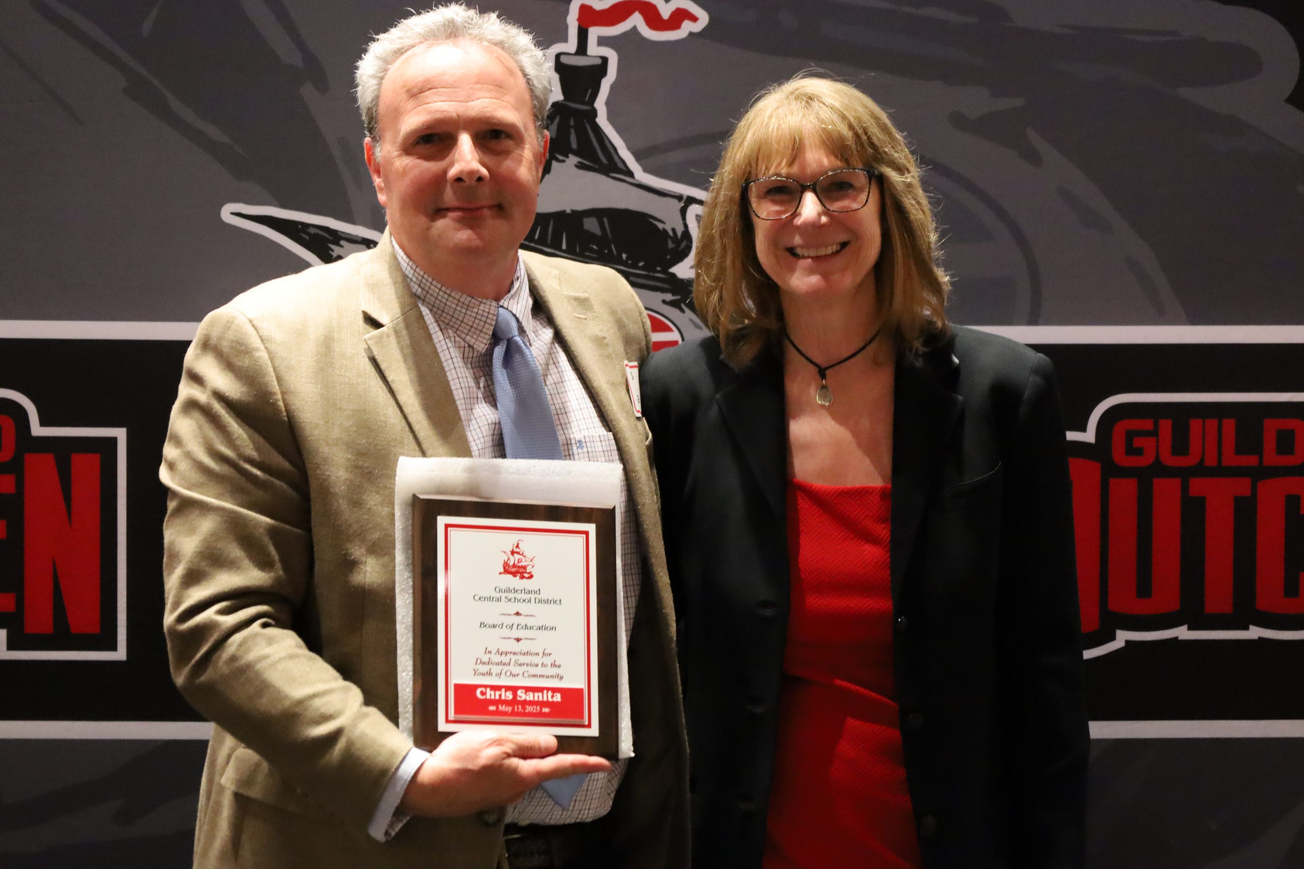 Two people stand in front of a backdrop. One is holding a plaque, both are smiling