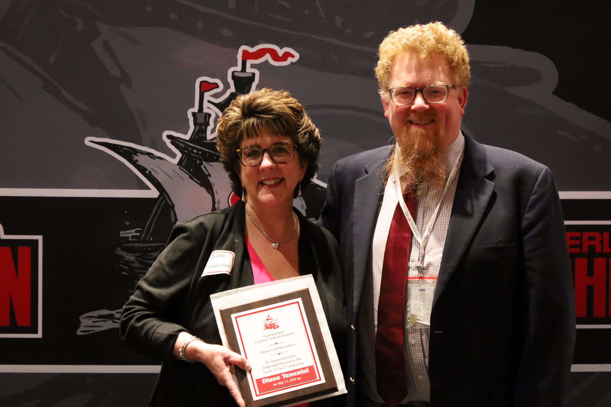 Two people stand in front of a backdrop. One is holding a plaque, both are smiling