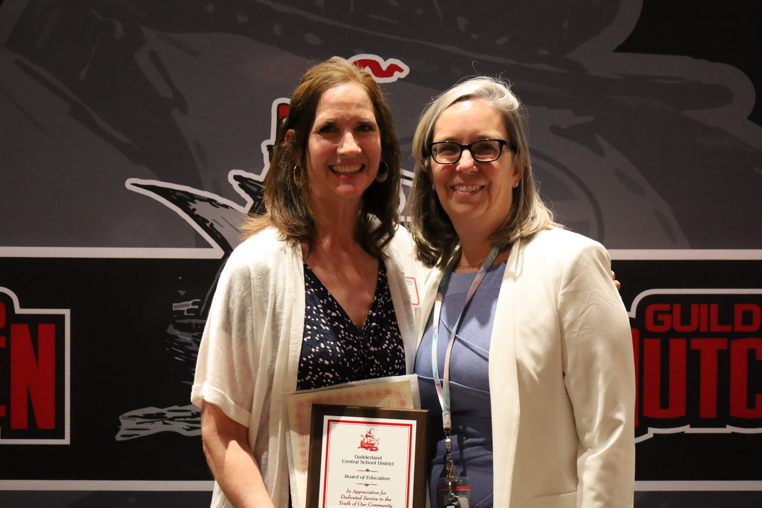 Two people stand in front of a backdrop. One is holding a plaque, both are smiling