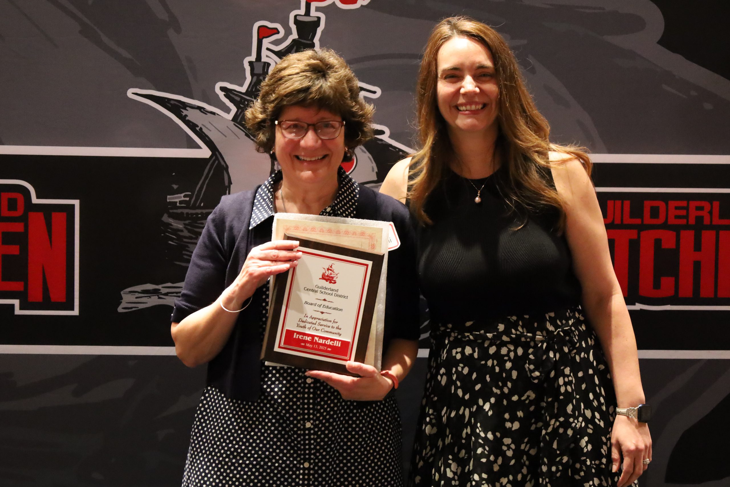 Two people stand in front of a backdrop. One is holding a plaque, both are smiling