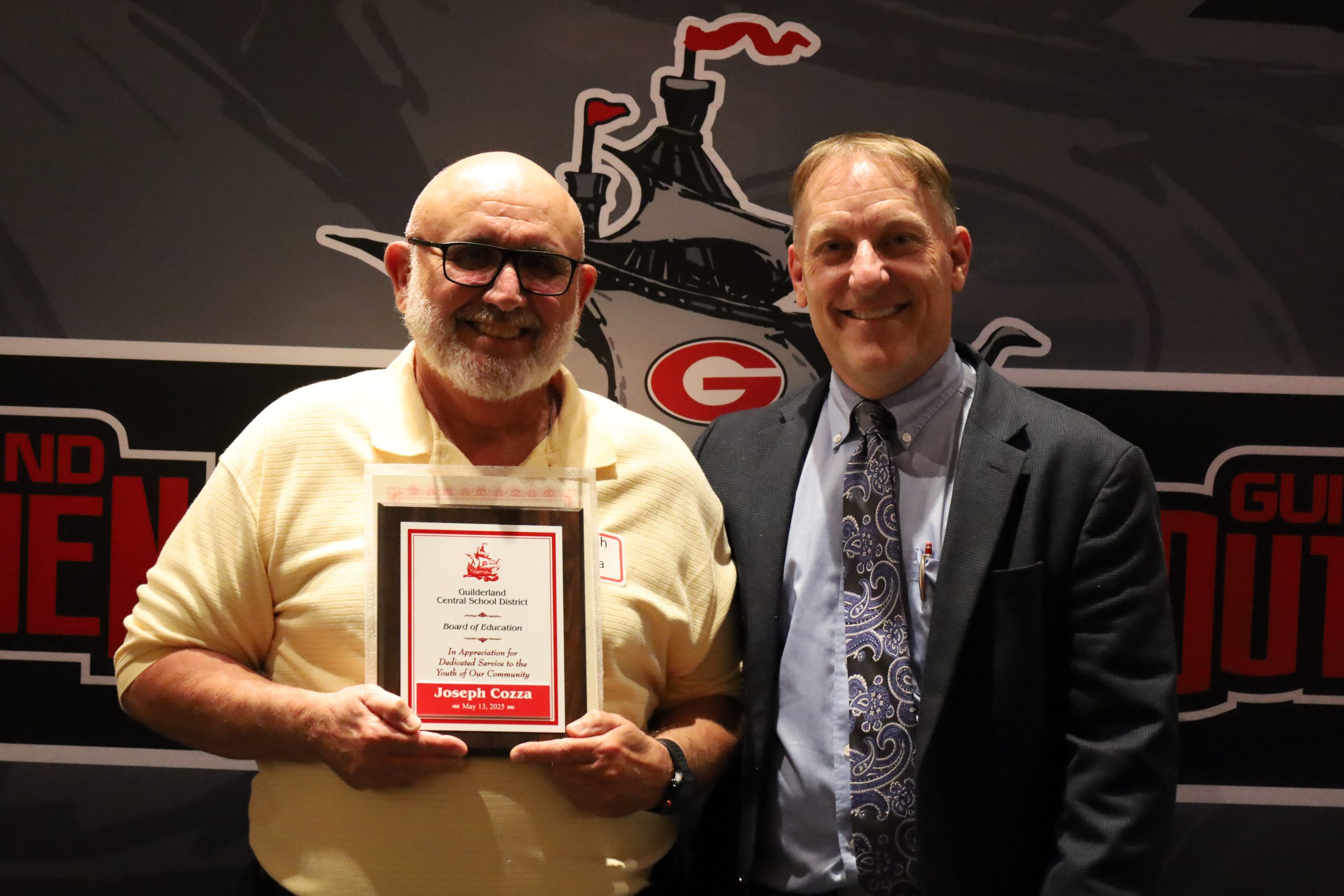 Two people stand in front of a backdrop. One is holding a plaque, both are smiling