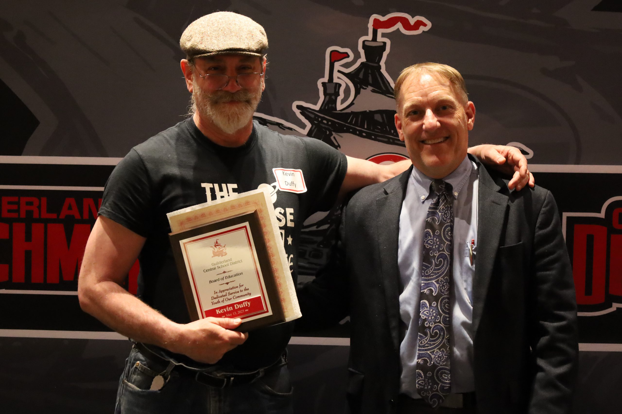Two people stand in front of a backdrop. One is holding a plaque, both are smiling