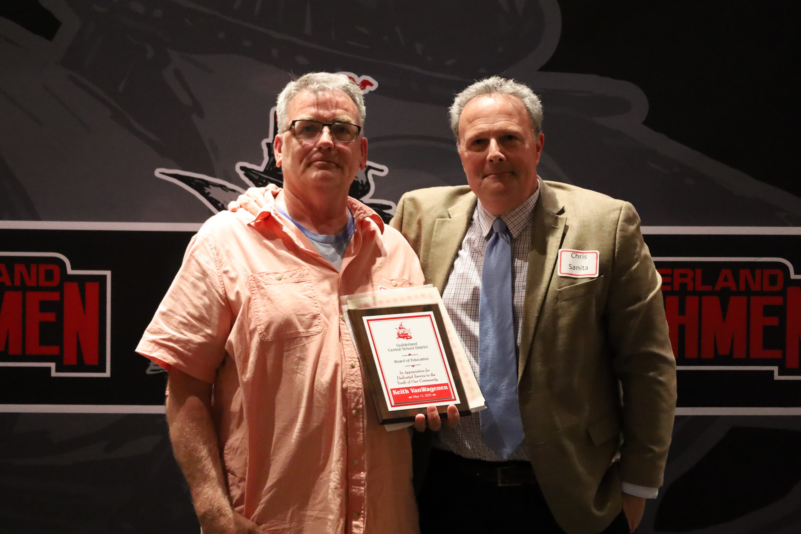 Two people stand in front of a backdrop. One is holding a plaque, both are smiling