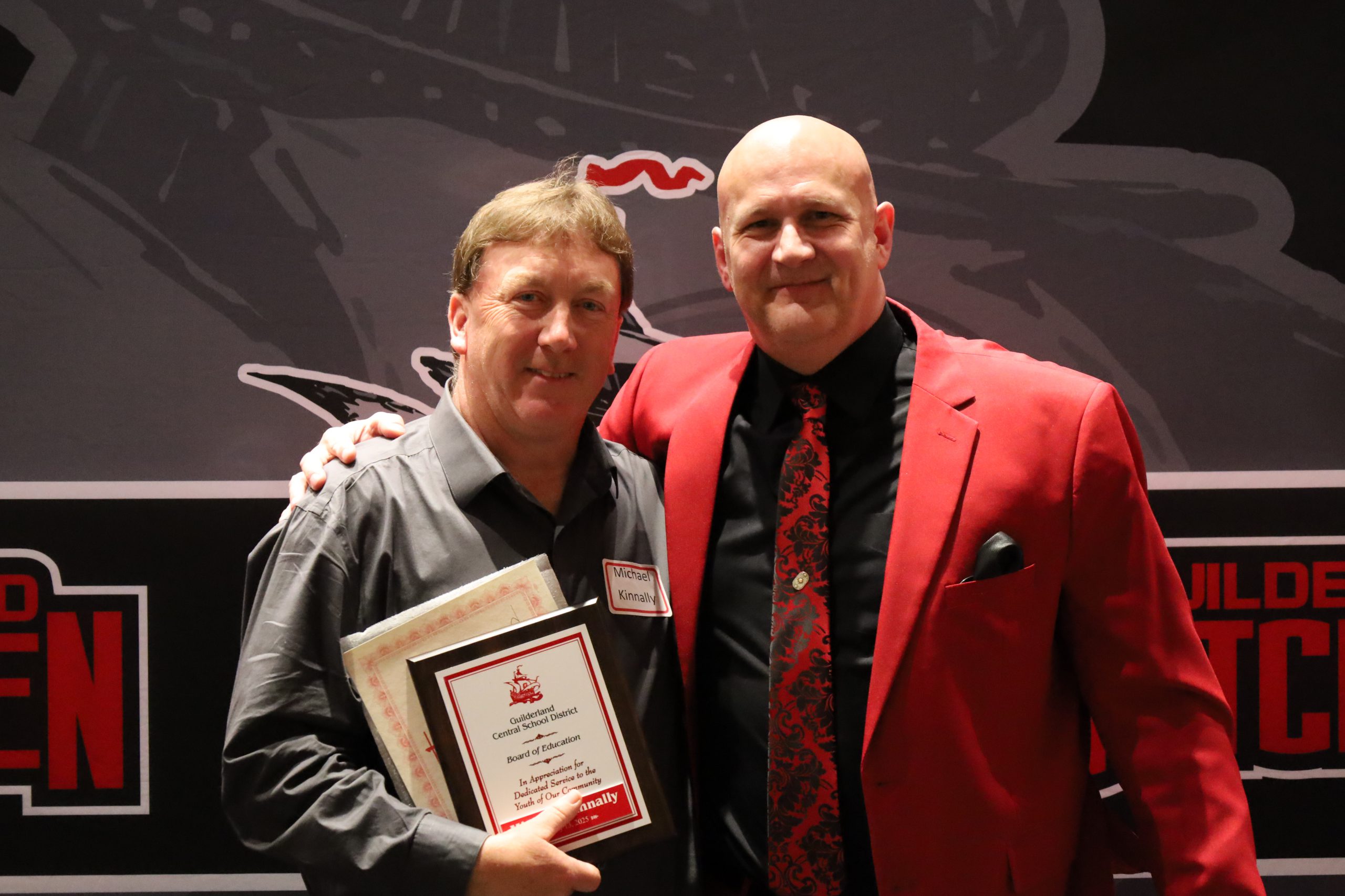 Two people stand in front of a backdrop. One is holding a plaque, both are smiling