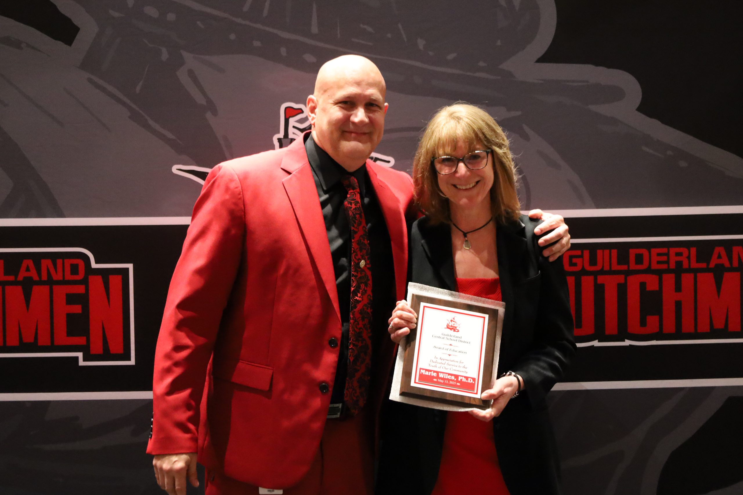 Two people stand in front of a backdrop. One is holding a plaque, both are smiling