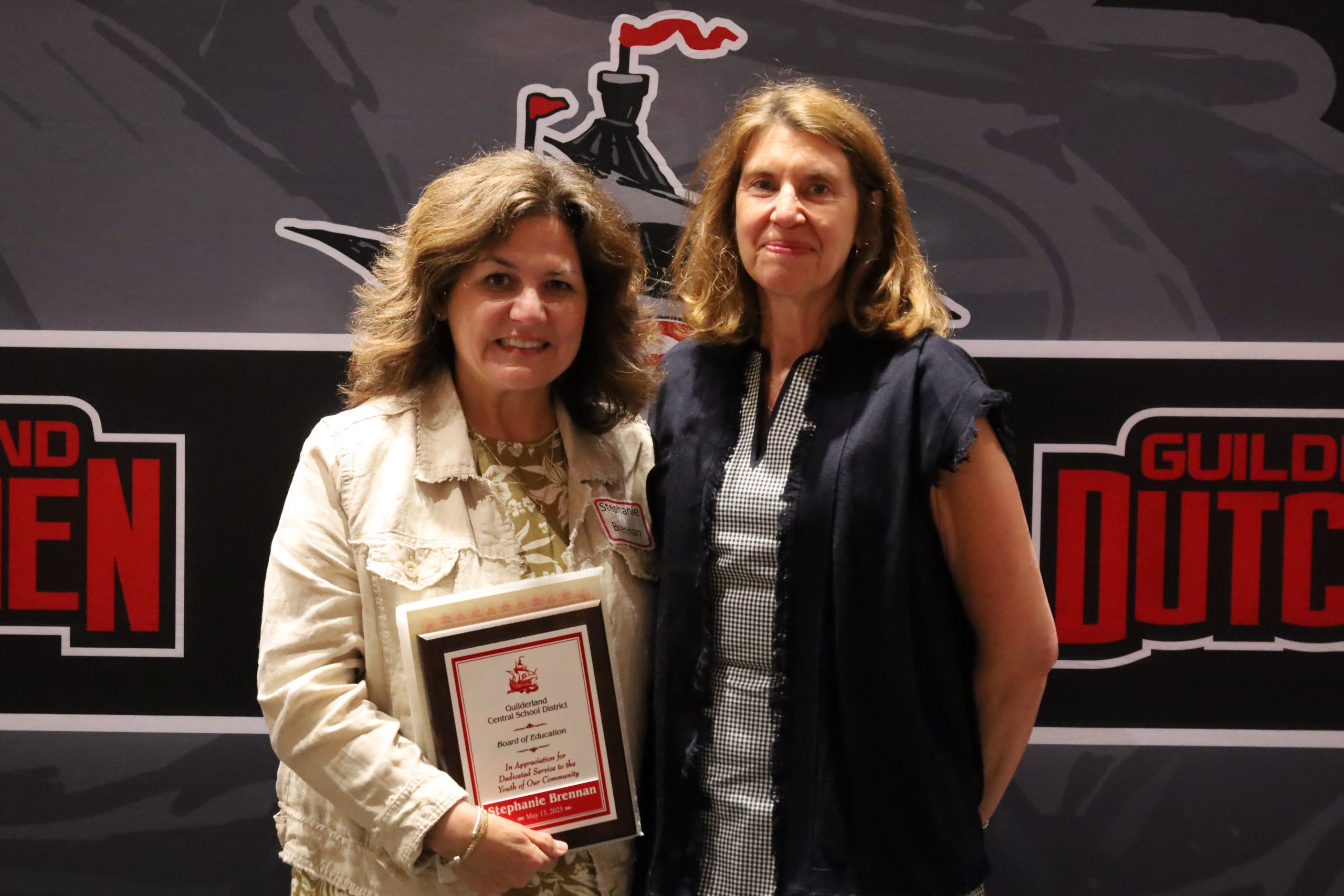Two people stand in front of a backdrop. One is holding a plaque, both are smiling