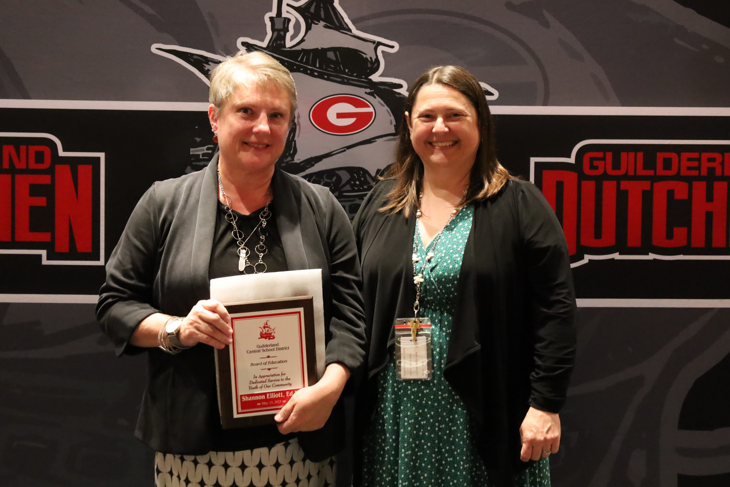 Two people stand in front of a backdrop. One is holding a plaque, both are smiling