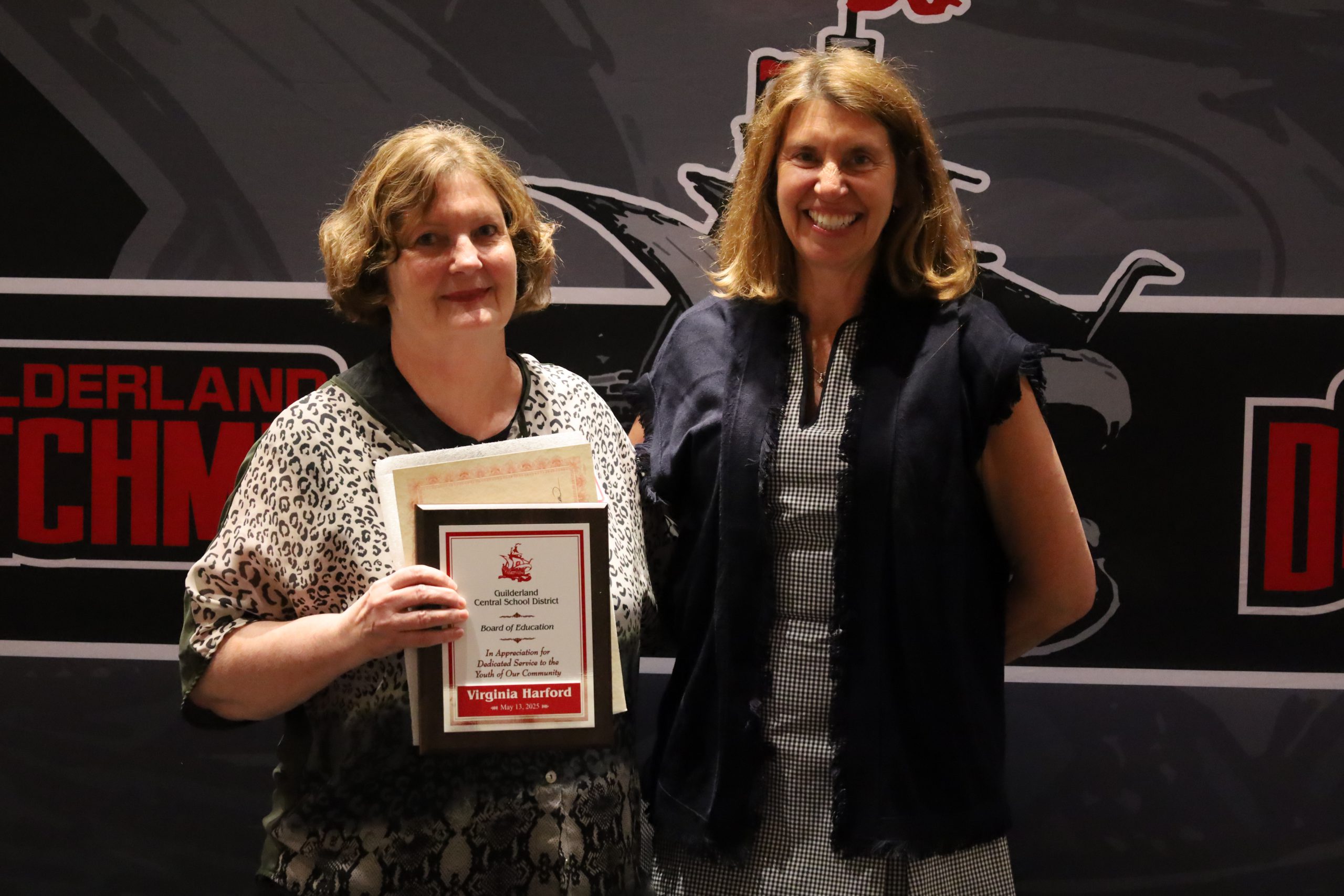 Two women stand in front of a backdrop. One is holding a plaque, both are smiling