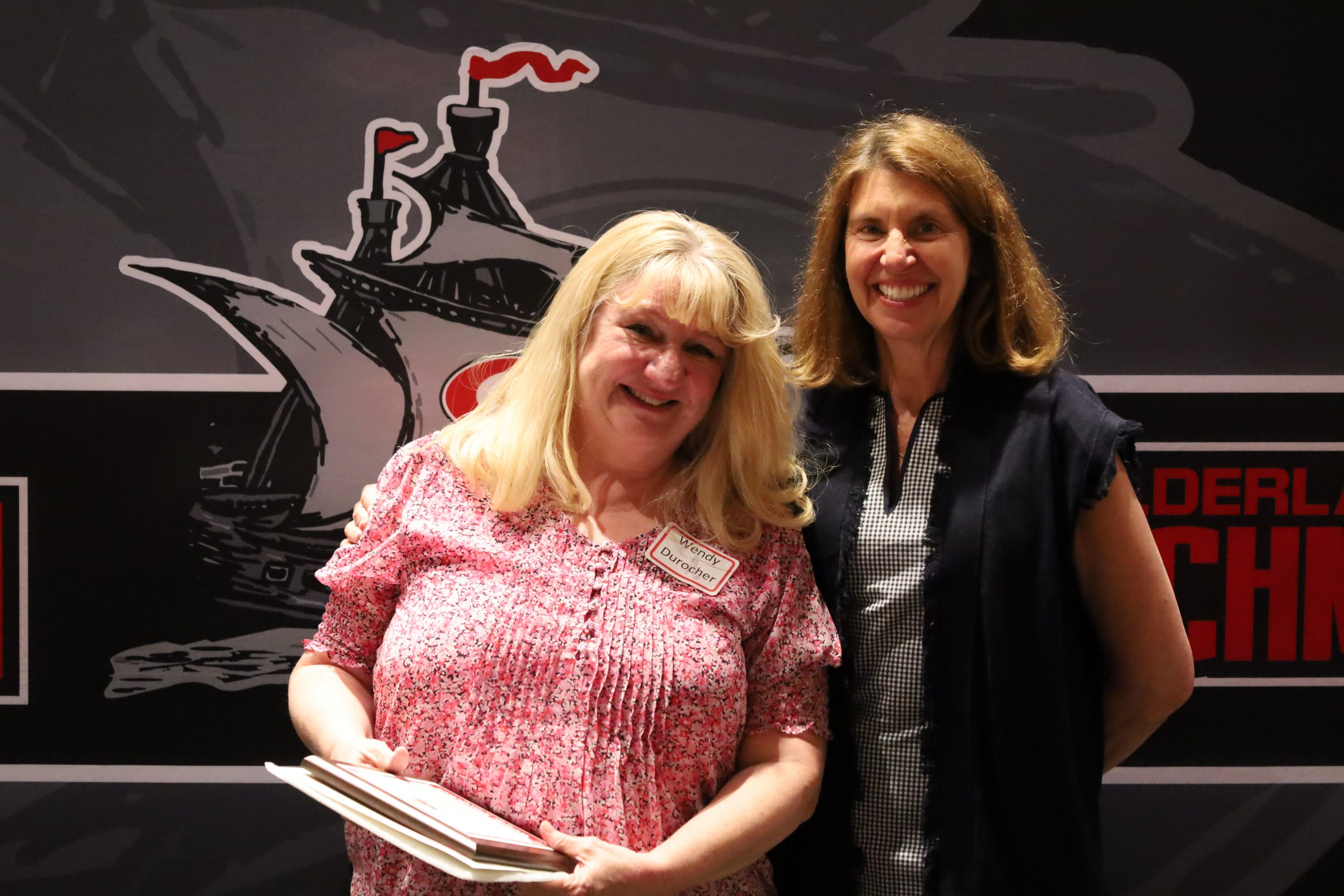 Two women stand in front of a backdrop. One is holding a plaque, both are smiling