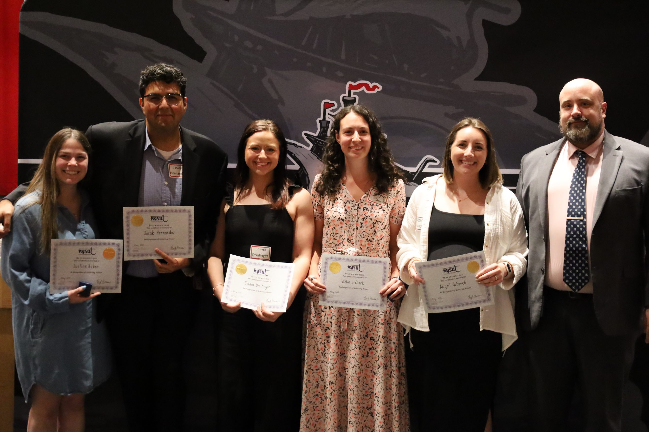 Six people stand in front of a backdrop, they are smiling. Five are holding certificates