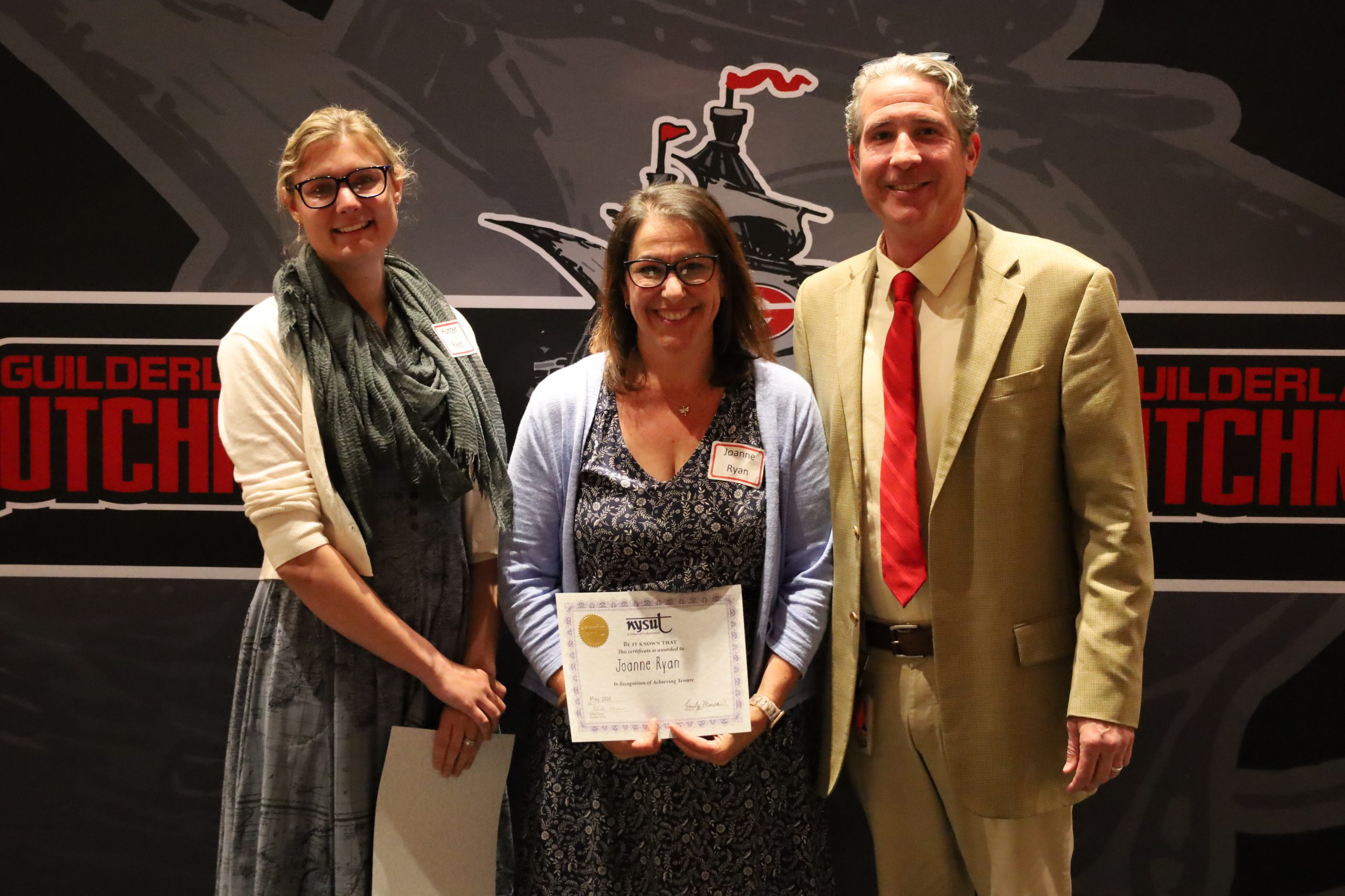 Three people stand in front of a backdrop, they are smiling. Two are holding certificates