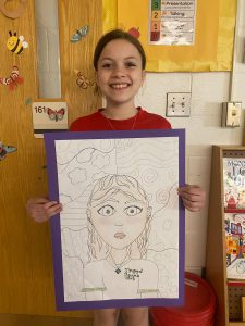 A young student stands in a classroom, holding a piece of artwork