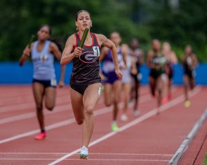 Female outdoor track athlete running on a red track with other runners behind her.