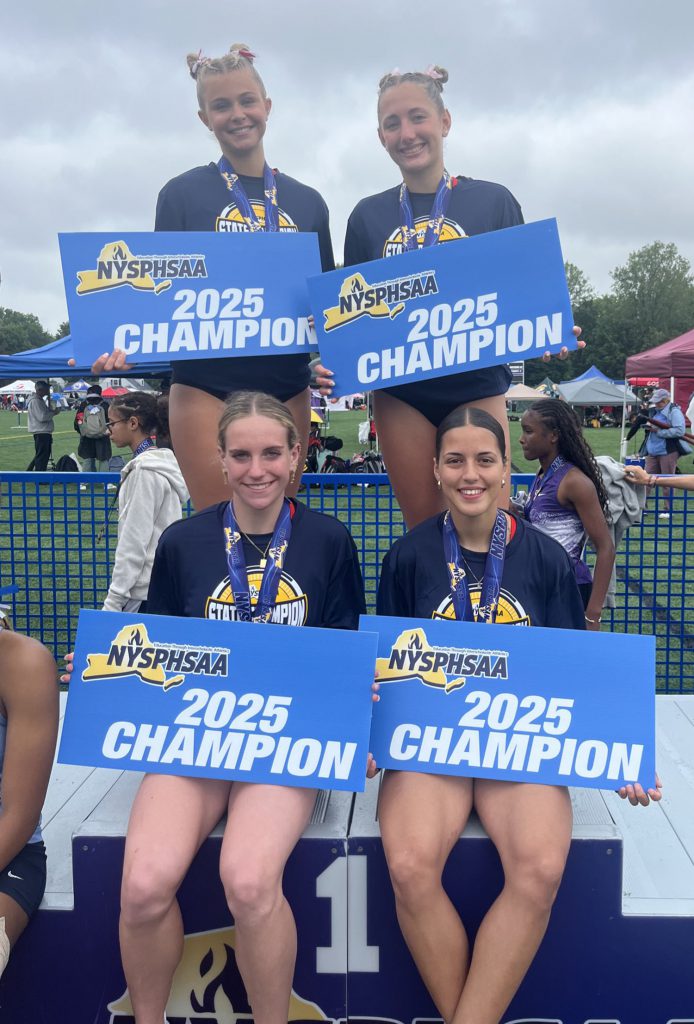 Four female athletes smiling for a photo with each holding a blue "NYSPHSAA 2025 Champion" sign in their hands. Two athletes are sitting in the front row, while the other two athletes stand behind them.