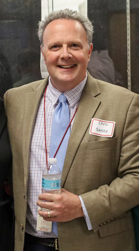 Man wearing a tan blazer with light blue tie and a nametag on the right side. He's smiling at the camera while holding a water bottle in his right hand.