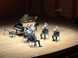 The photo is taken from above. It's of five student-musicians seated on a stage in a semi-circle, playing brass instruments