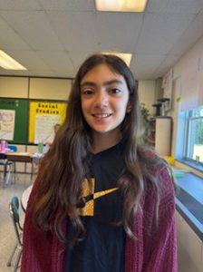 A student stands in a classroom, smiling, wearing a dark t-shirt and a raspberry colored sweater