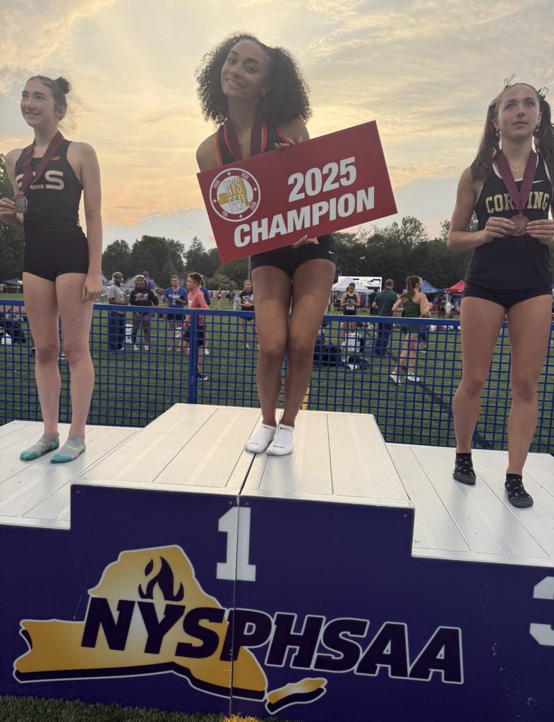 Three athletes standing on the NYSPHSAA podium that has first place in the middle, second place on the left and third place on the right. The GHS female athlete is standing on the first place spot holding a red "2025 Champion" sign in her hands.