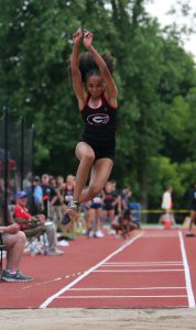 Female track athlete jumping in mid-air with the red track in the background.