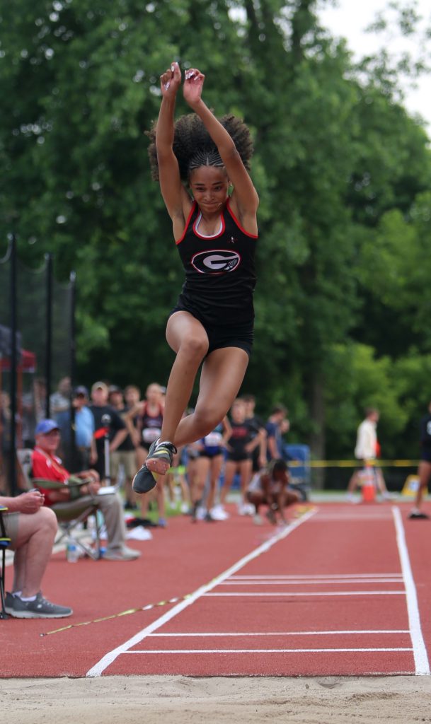 Female track athlete jumping in mid-air with the red track in the background.