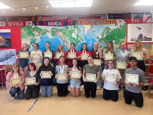 18 students are in a classroom, in two rows. The front row is kneeling, the back row is standing. All are holding certificates, some are wearing medals.