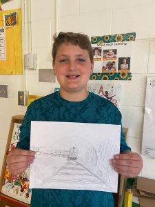 A student stands in a classroom and is displaying a piece of artwork