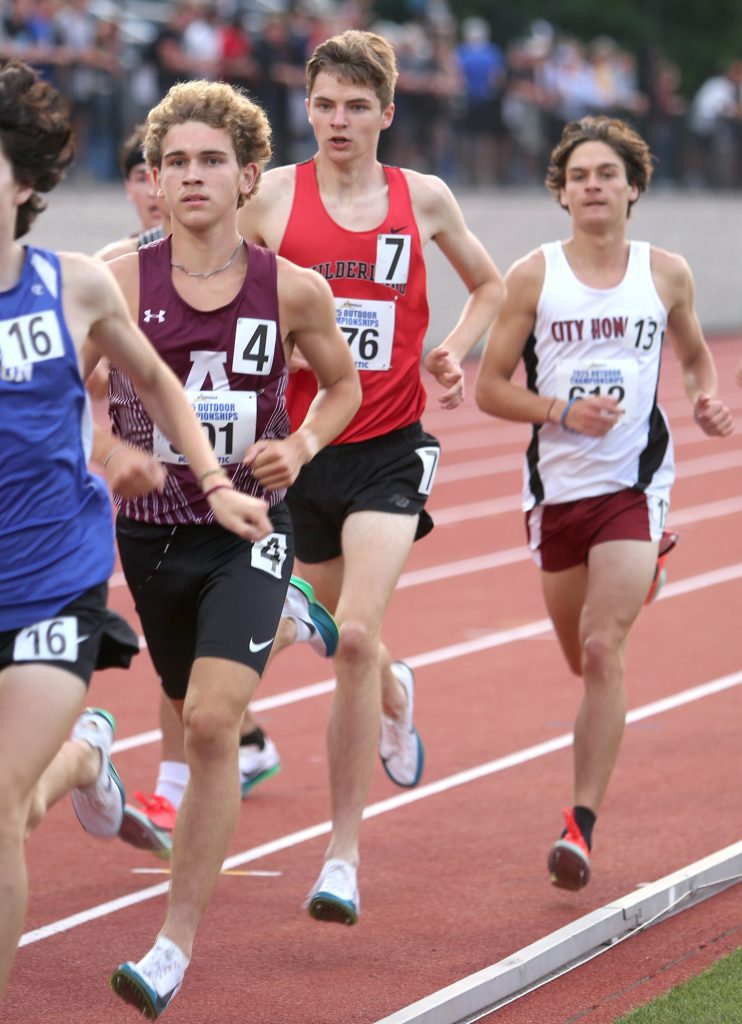 Three male athletes running on the track in their uniforms.