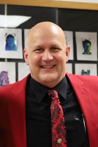 A man wearing a red suit jacket, black shirt and red and black tie smiles at the camera