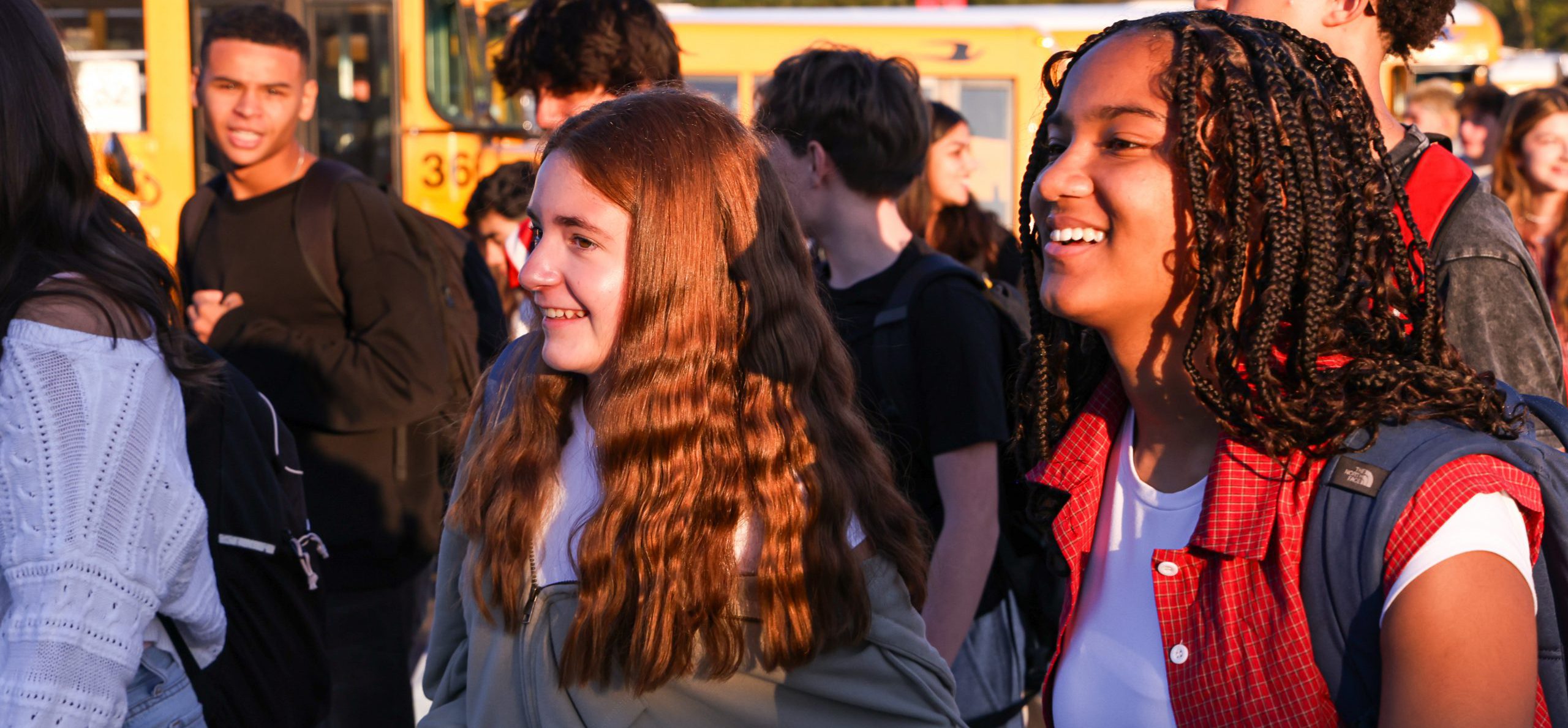 High school students smiling on the first day of school with buses in the background.
