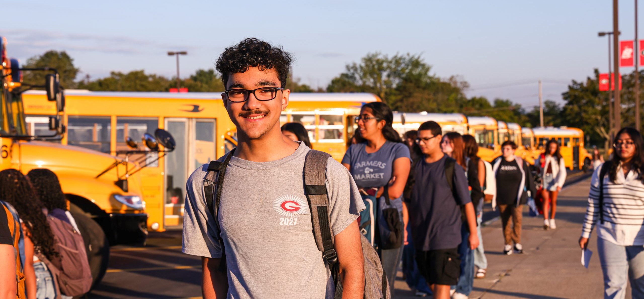 Student smiling at camera on first day of school with buses in the background.