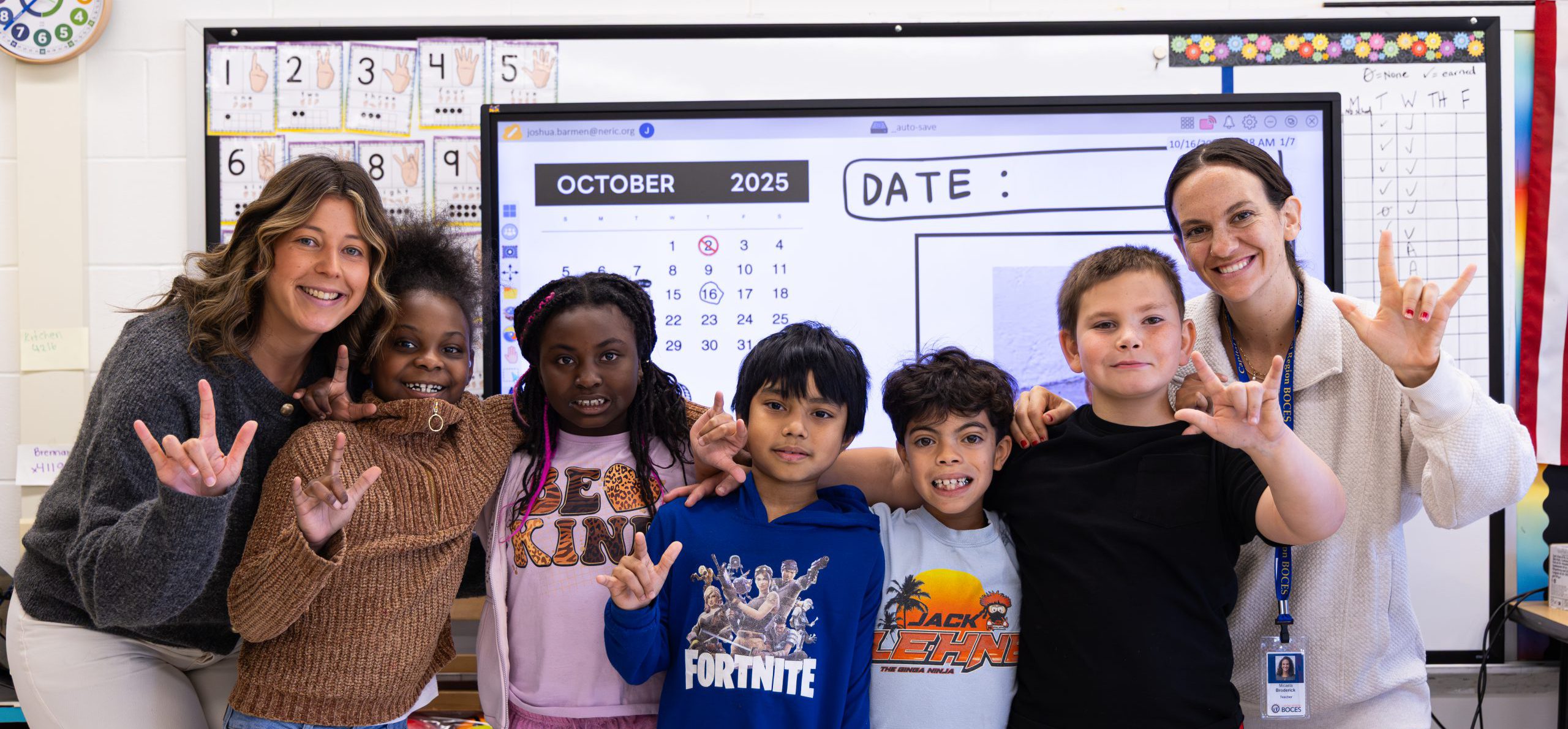 WES DHH staff and students smiling and signing "I love you" at the camera. They are standing in a classroom.