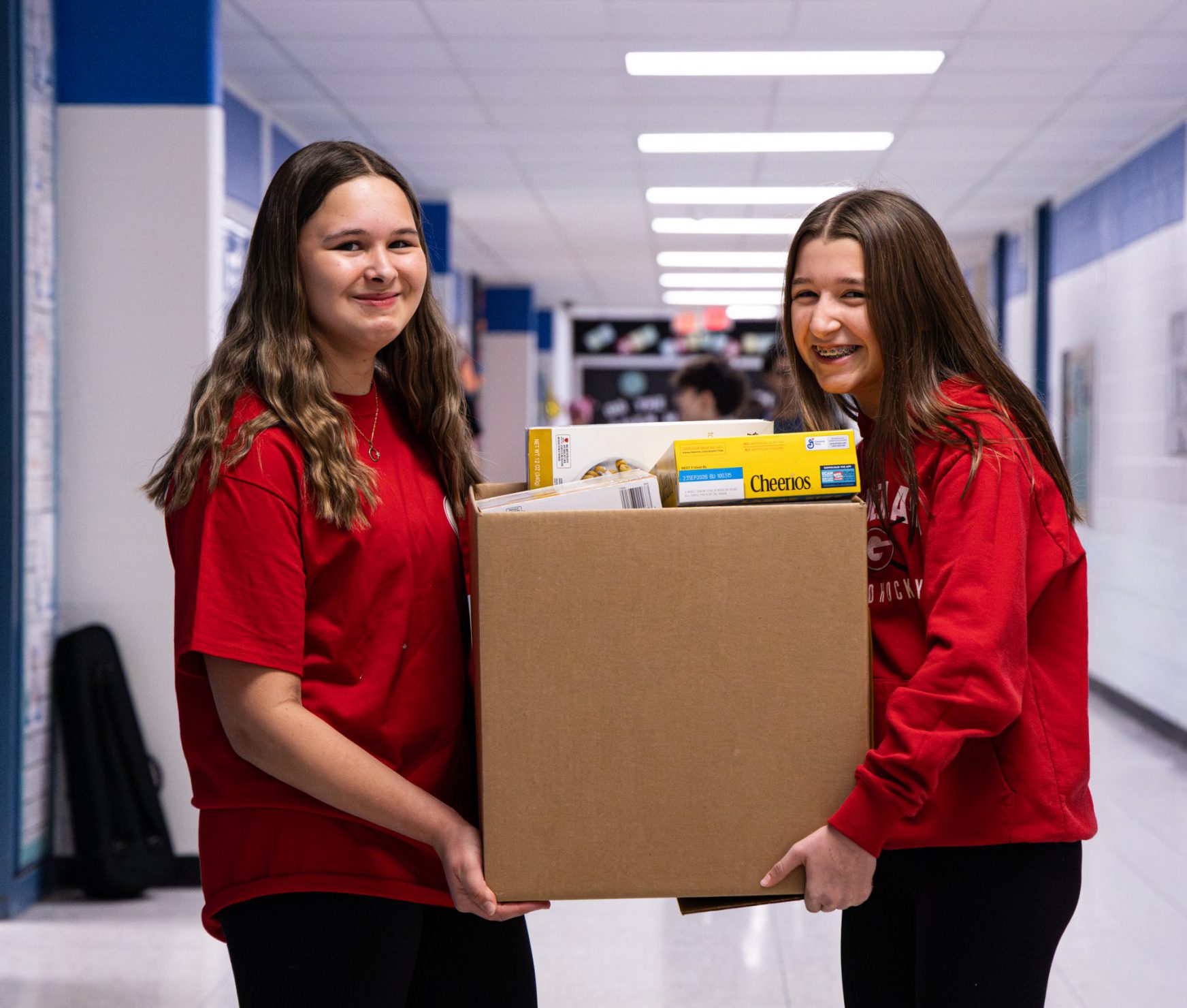 Two FMS students smiling in the halls. They are holding a box filled with cereal boxes in it to be donated to the food pantry.