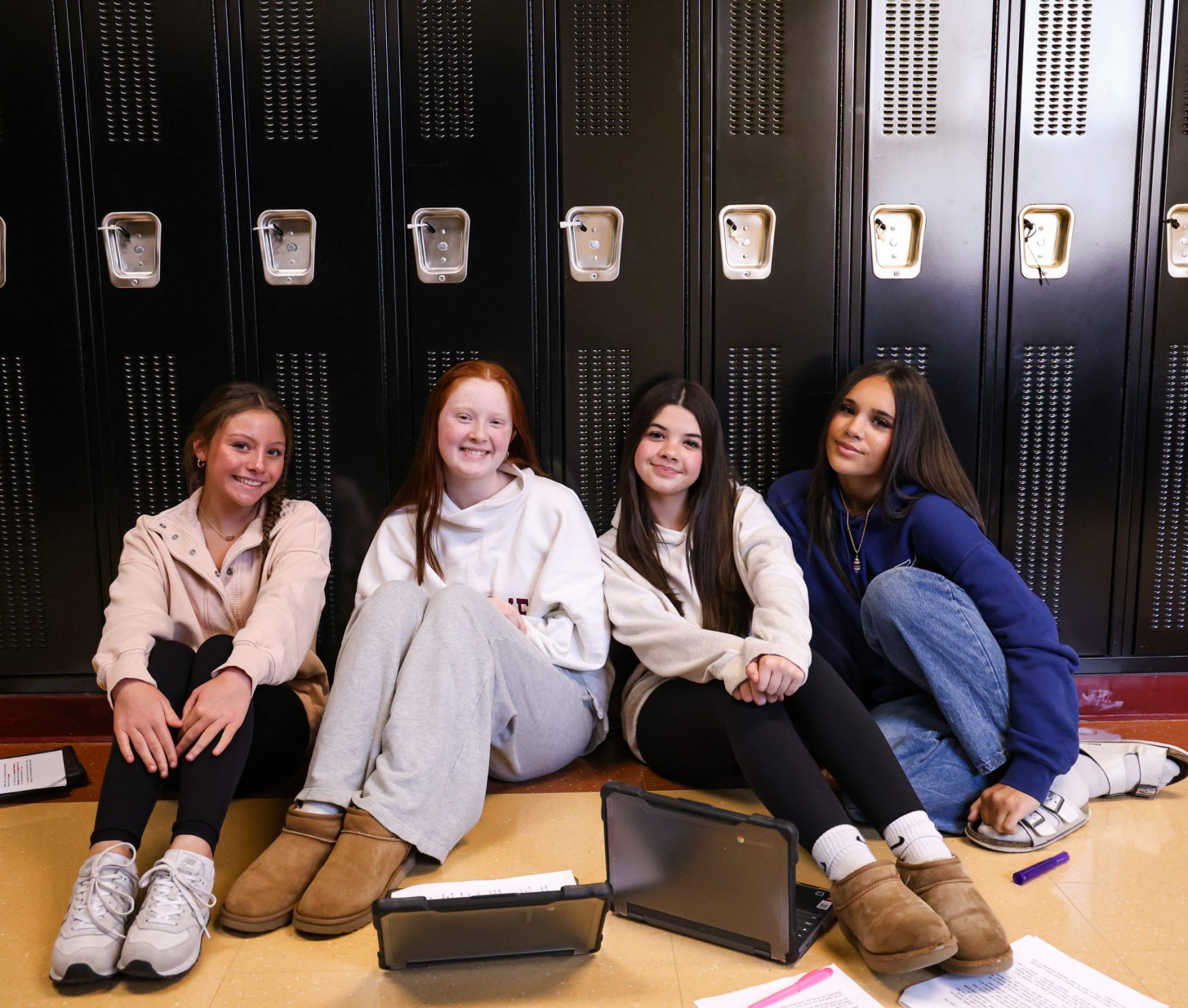 Four GHS students smiling in hallway. They are sitting on the ground with lockers behind them.