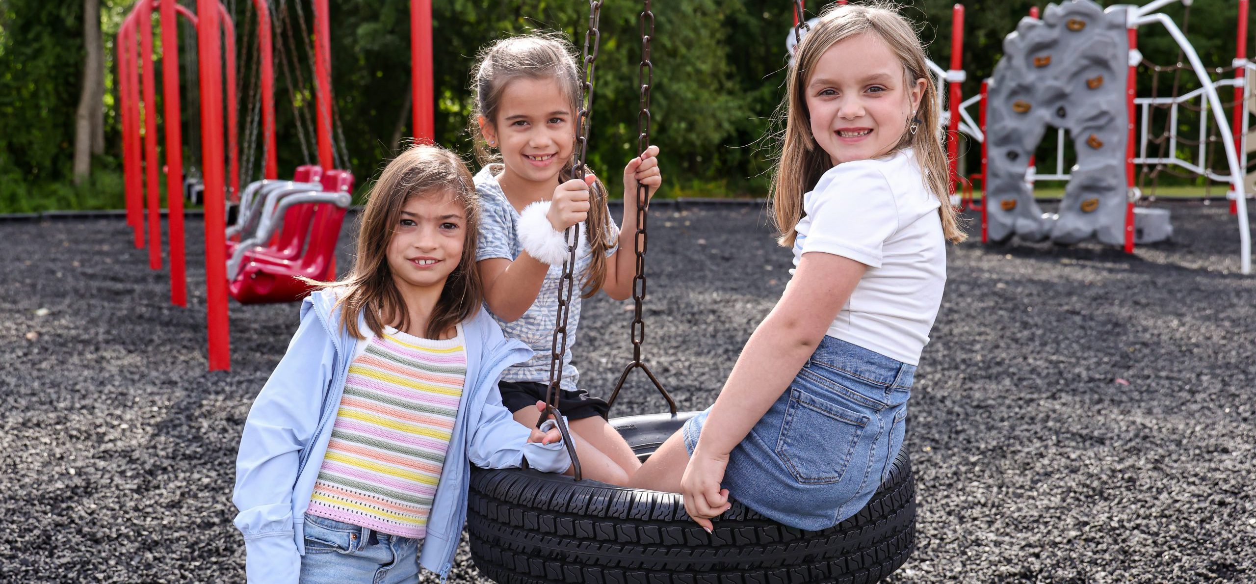 Three children smile at the camera on a tire swing on a playground